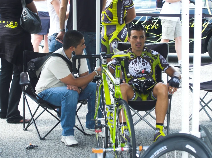a man sits on a folding chair beside another man with a bike in their hands