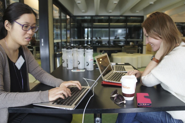 two women are sitting at a table with laptops