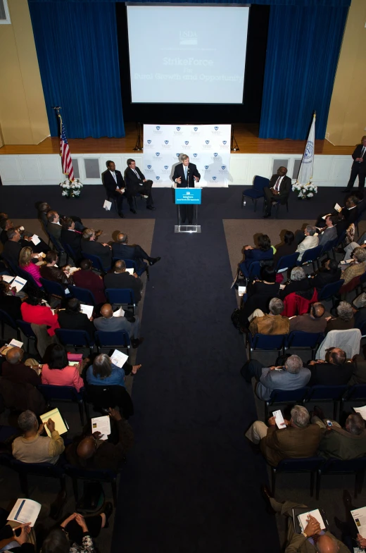 two people speaking at a podium with several seated up people in the background