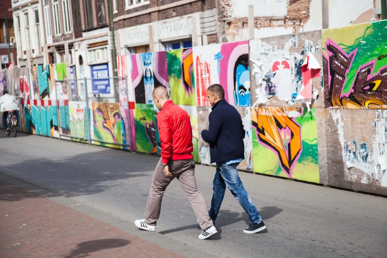 two people are walking on a sidewalk by a wall with graffiti