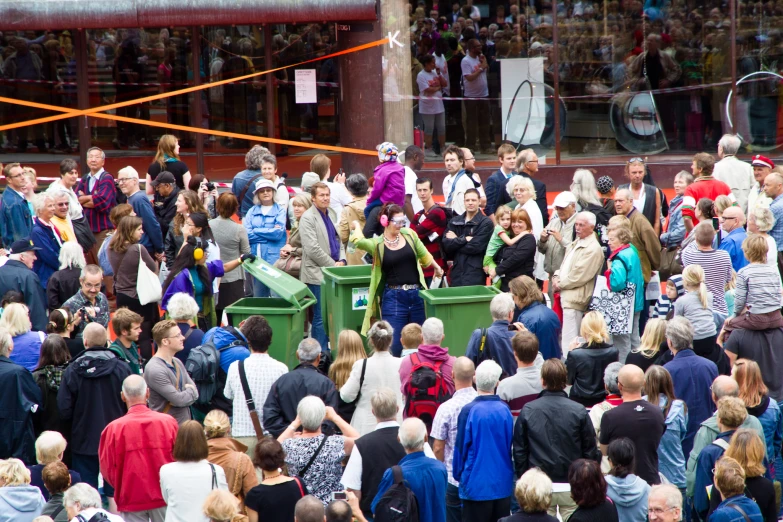 a crowd is seen standing around a green garbage container