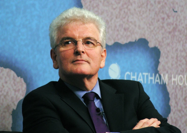 an older man wearing glasses and a suit in front of a wall