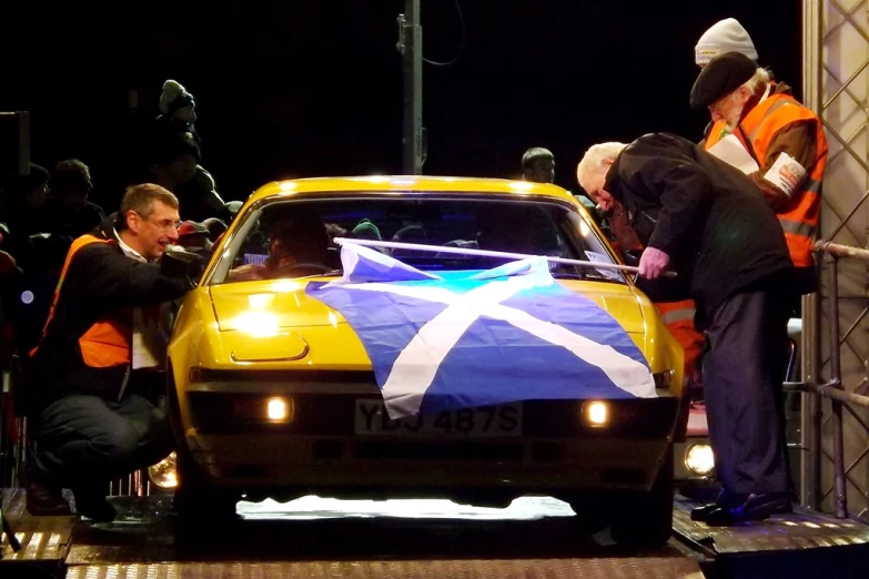 several people next to an automobile with a flag painted on the hood