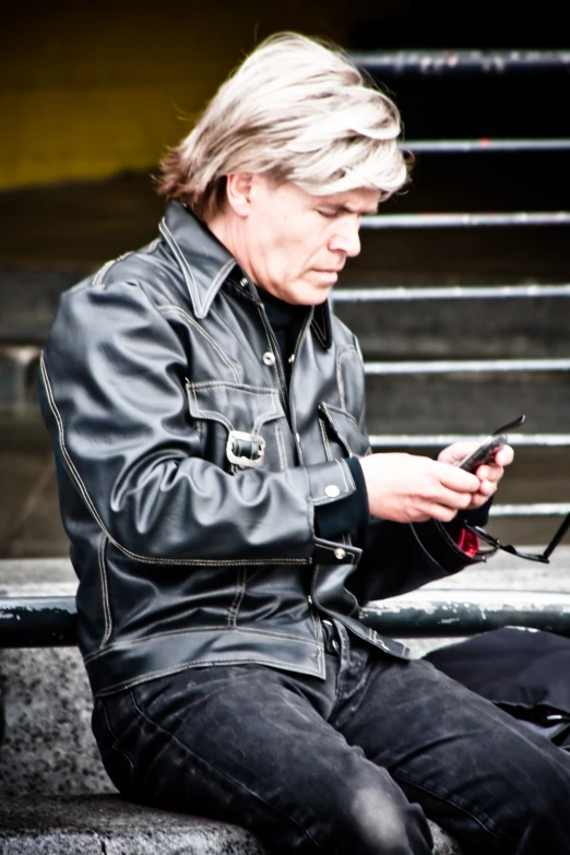 a man in black jacket sitting on a ledge using his phone