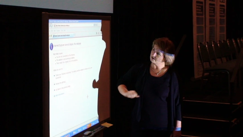 a woman is speaking in front of a large screen