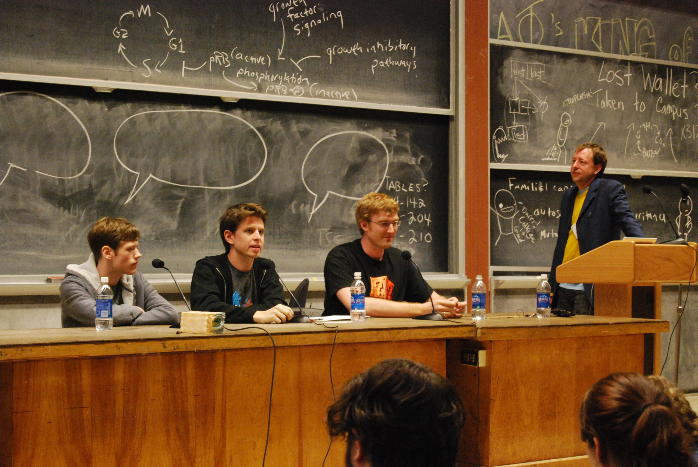 people sit in front of a classroom while two other men speak