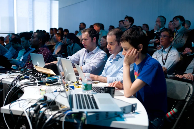 three men sitting in front of laptop computers with people watching them