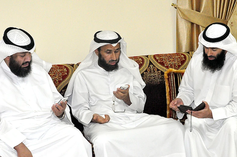 four men are sitting on a couch looking at cell phones