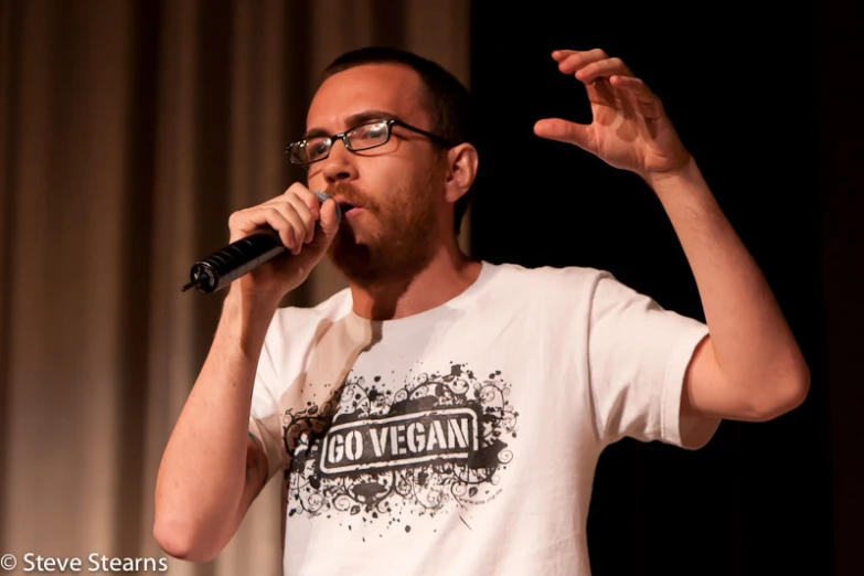 a man with glasses holds his hand out for the camera as he speaks into a microphone