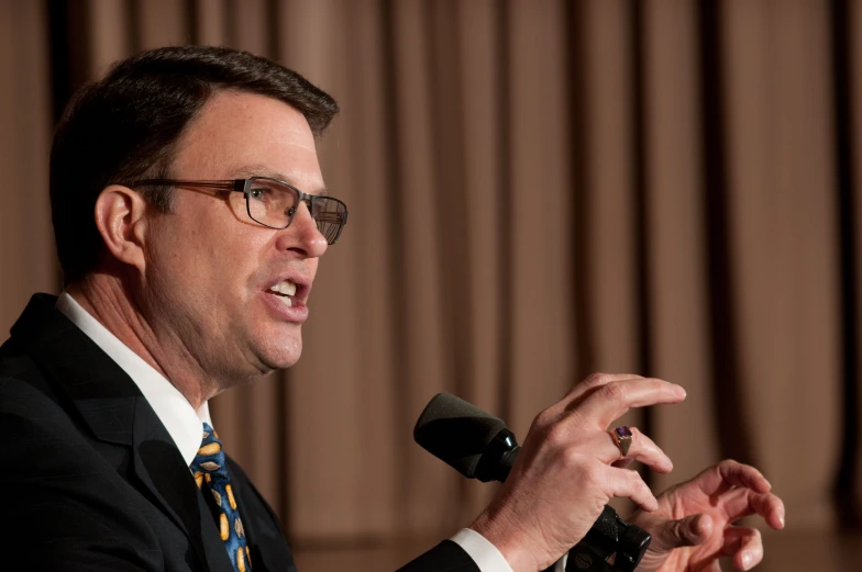 man talking at podium holding microphone and standing in front of curtains