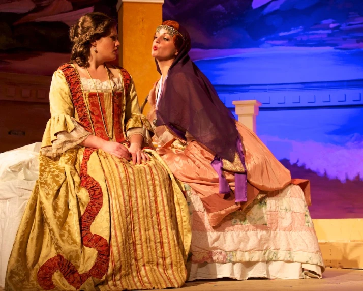 two women sit on a bed during a performance