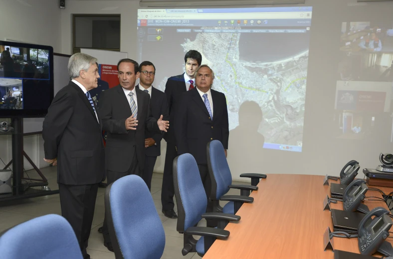 men in suits standing near a desk with a large screen