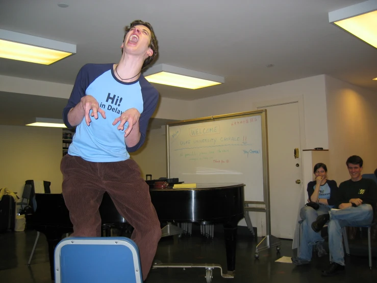 a young man stands on a chair in a classroom while a group of people look on