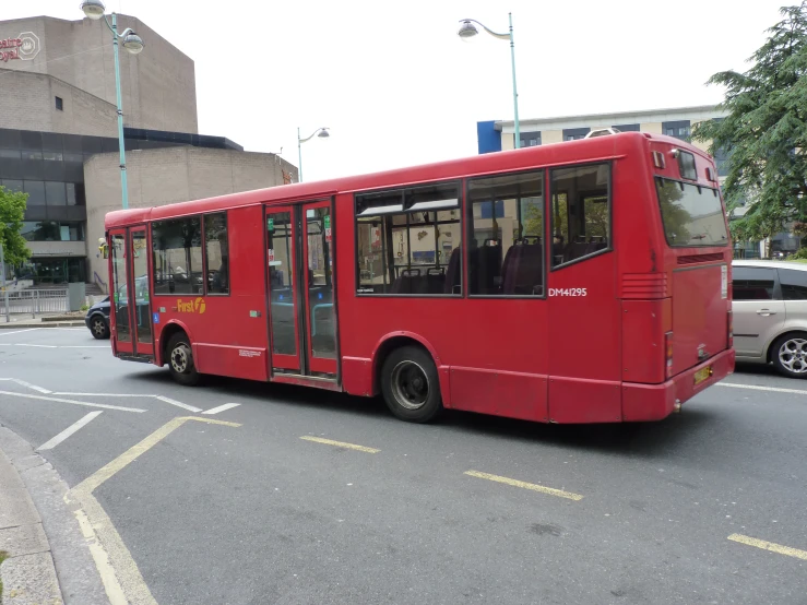 a red bus going down the road with a large building in the background