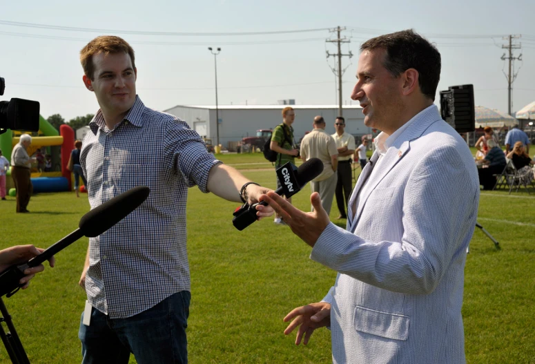 a man talks to a man holding a microphone in front of an outdoor crowd