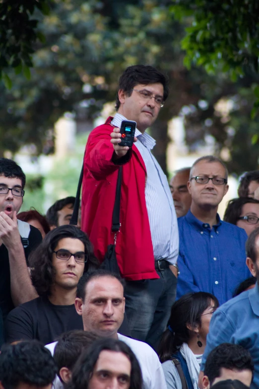 there is a male standing on the back of a crowd with a cellphone in his hand
