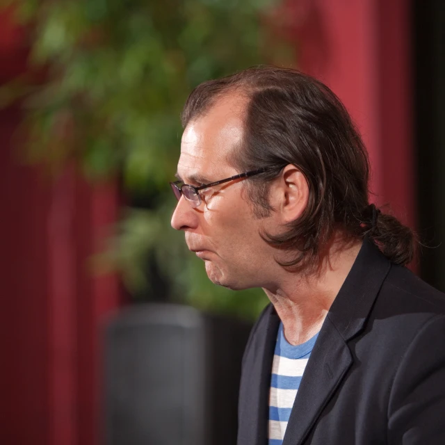 a man wearing glasses and a jacket standing in front of a tv