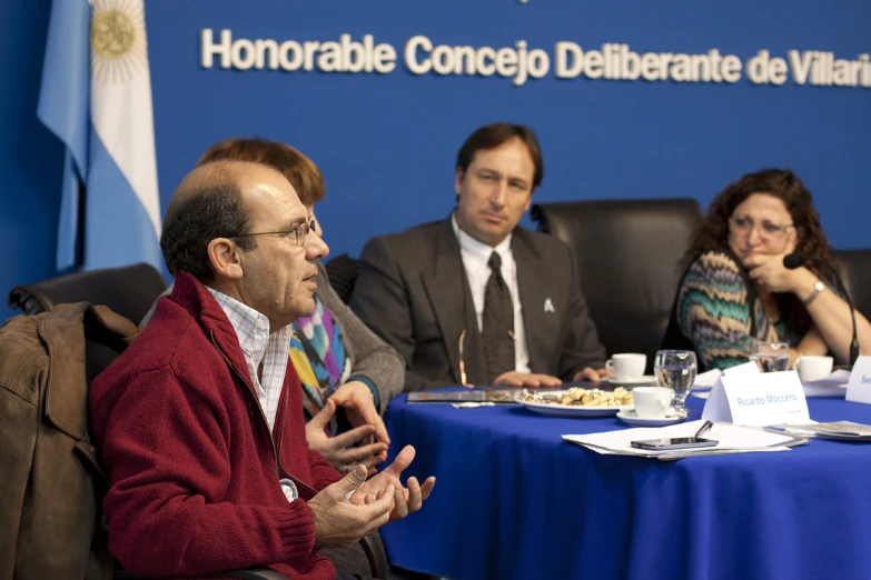 three men and two women sit at a table in front of a blue sign