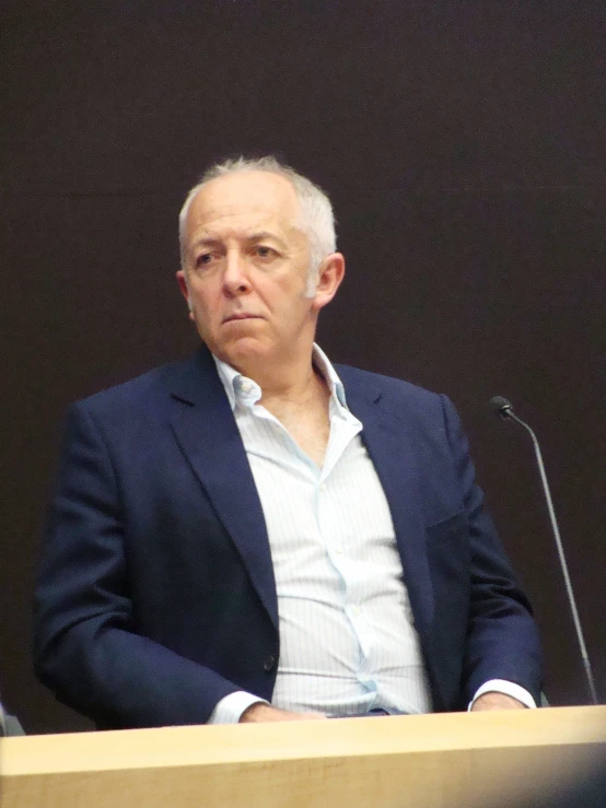 an older man sitting behind a podium in front of microphones