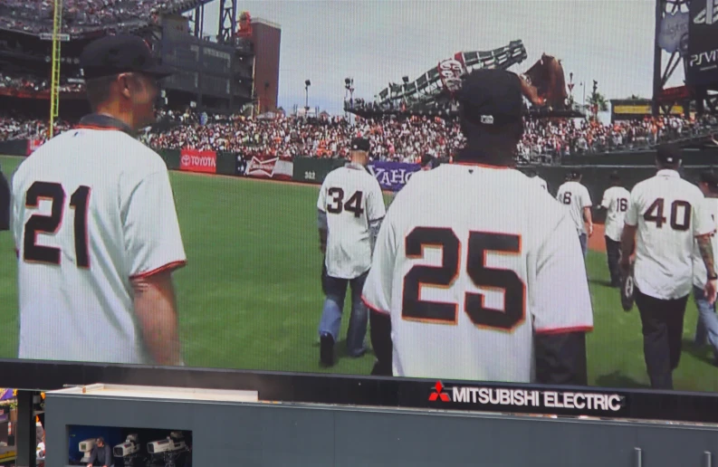 baseball players are standing in a field, with a television screen behind them