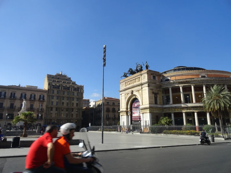 two people riding mopeds down the street near a building