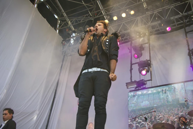 a man in black pants stands on stage and sings into microphones