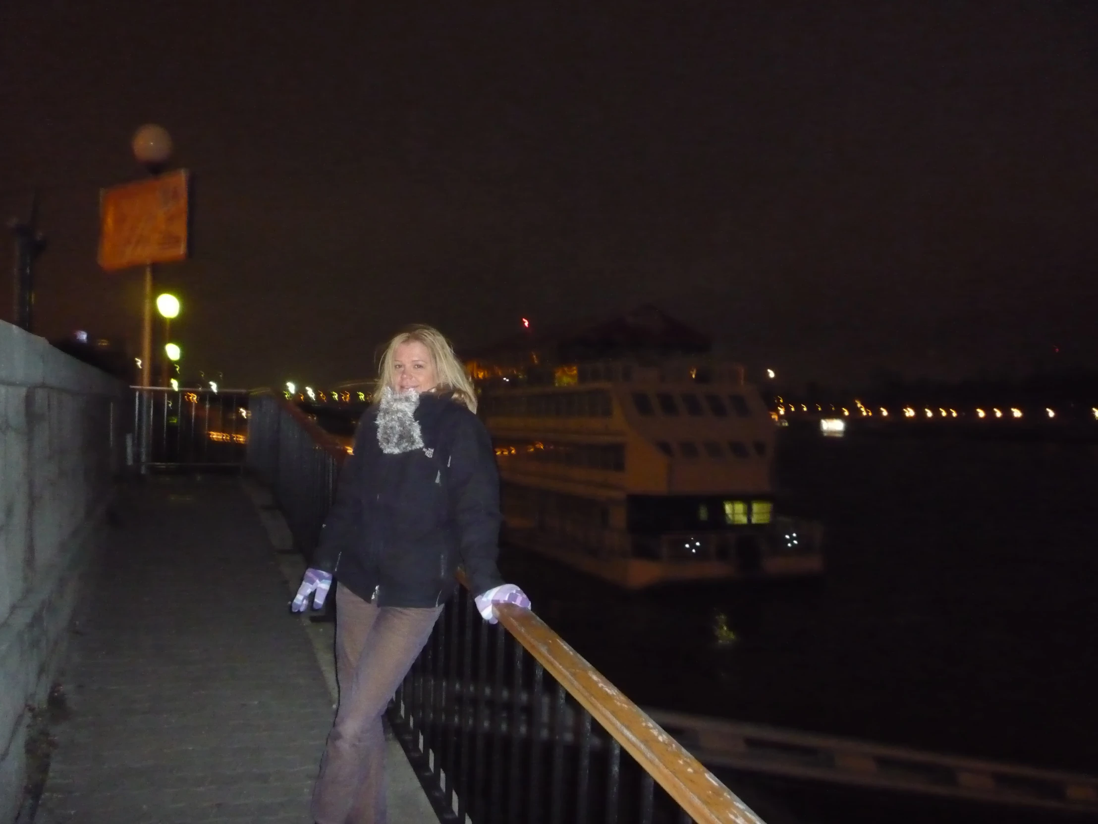 woman standing on a boardwalk by water with city lights at night