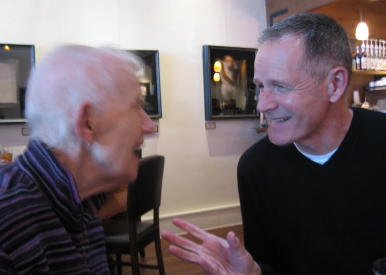 an older man sitting at a table talking to another older man