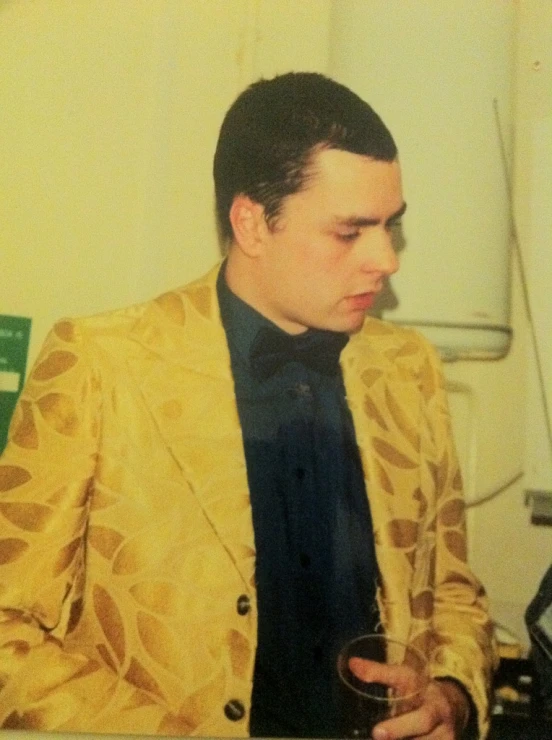 a man dressed in a yellow suit sitting and holding a glass
