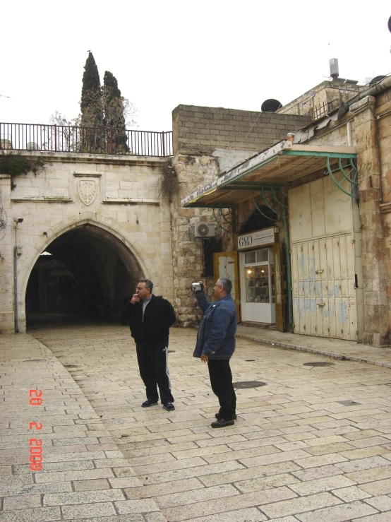 two men standing together on cobblestone area in front of a building