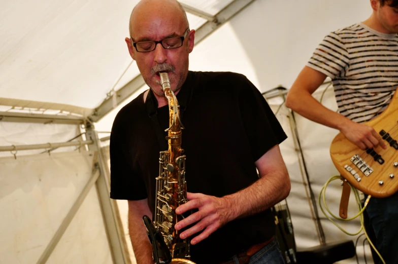 a man playing a saxophone and wearing glasses while another man plays a guitar behind him