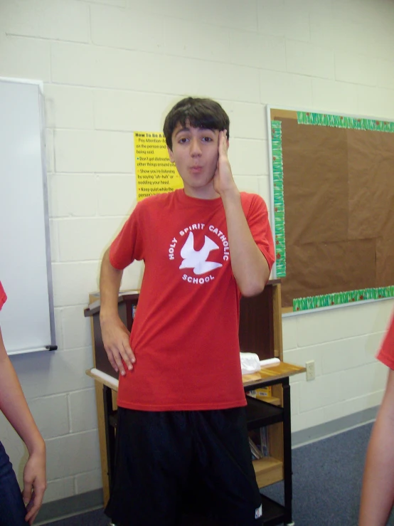 boy holds up one hand to his face while standing in a classroom