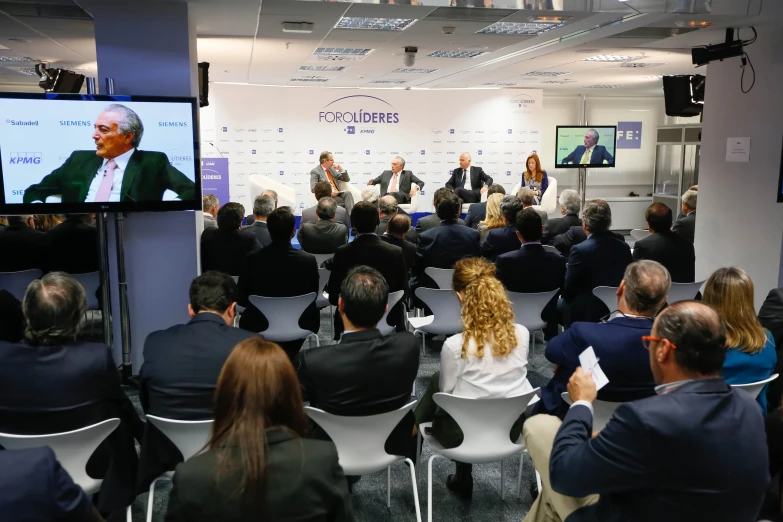 people sitting in chairs watching men on a video screen