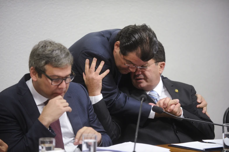 two men sitting at a table one is touching the other's head