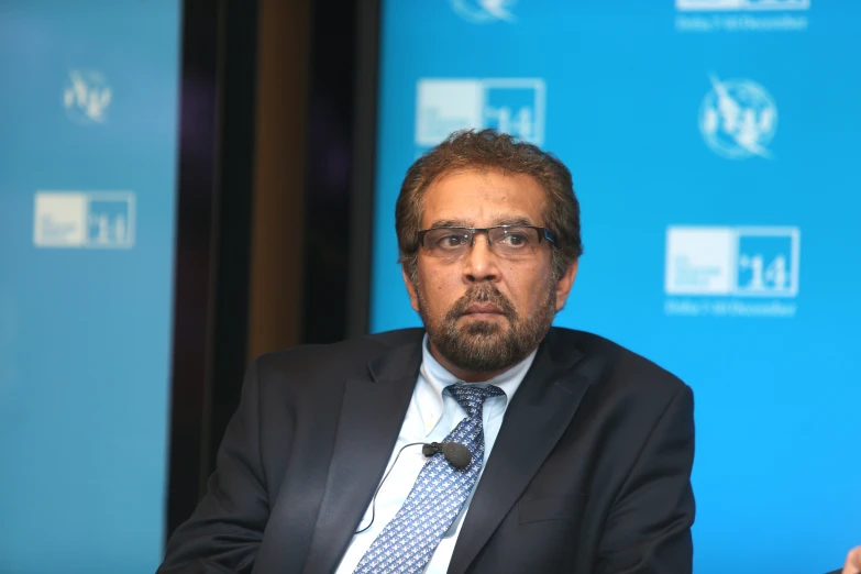 a man in a business suit and tie sits in front of a blue screen