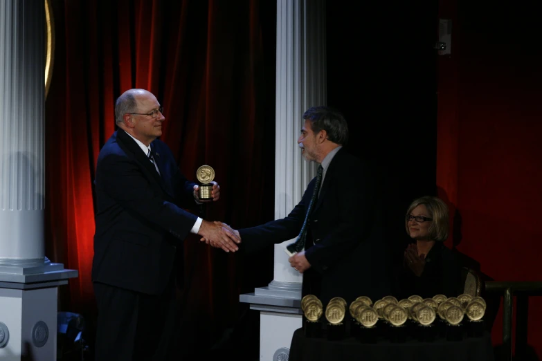 two men shake hands in front of red curtains