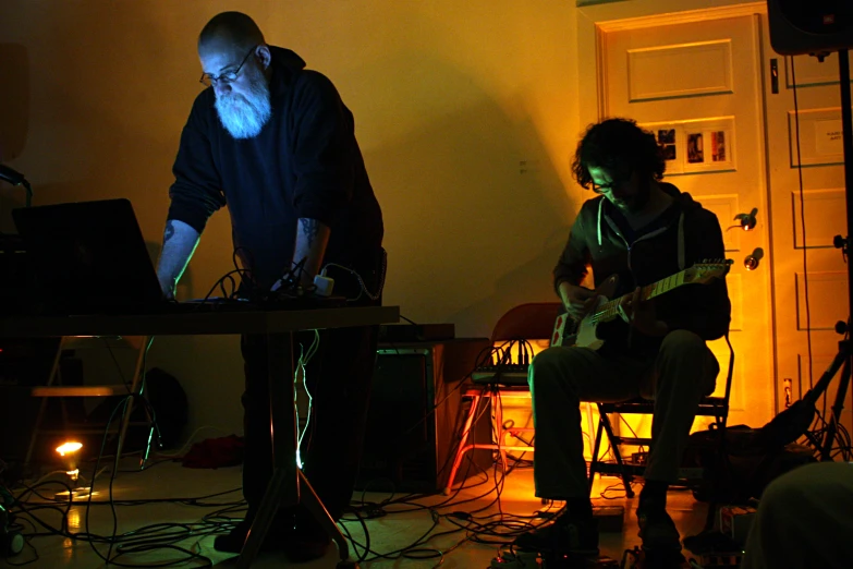 a man with a beard standing in front of another man playing guitar