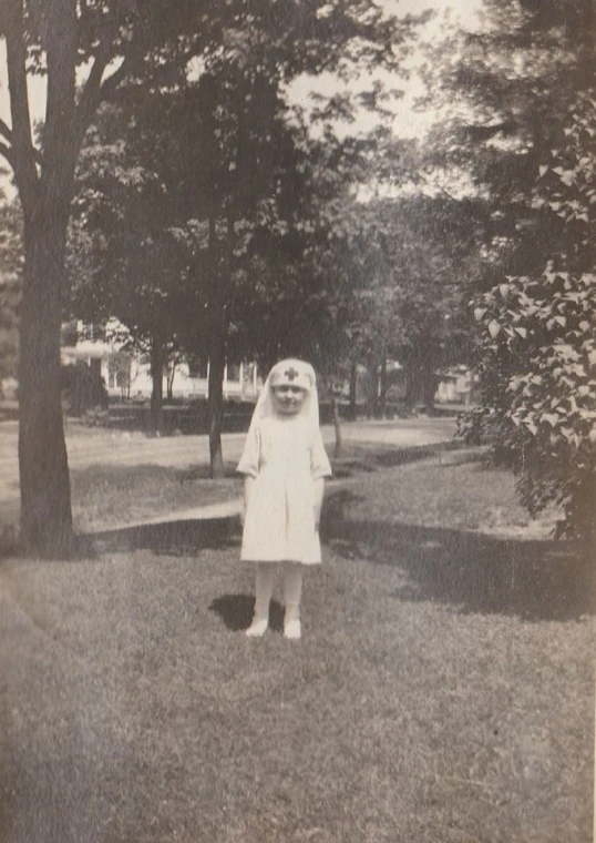 an old pograph of a woman in a white dress standing in the grass in front of a tree