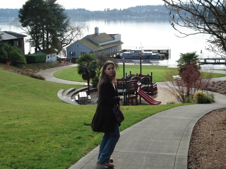 a woman in a black coat and jeans standing on the edge of a path next to a river