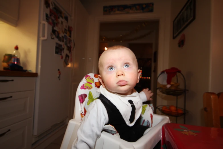 a baby sitting in a high chair at the kitchen table looking up