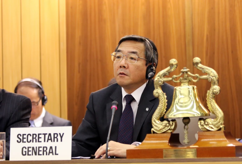 a man in suit and tie sitting at a desk with microphones