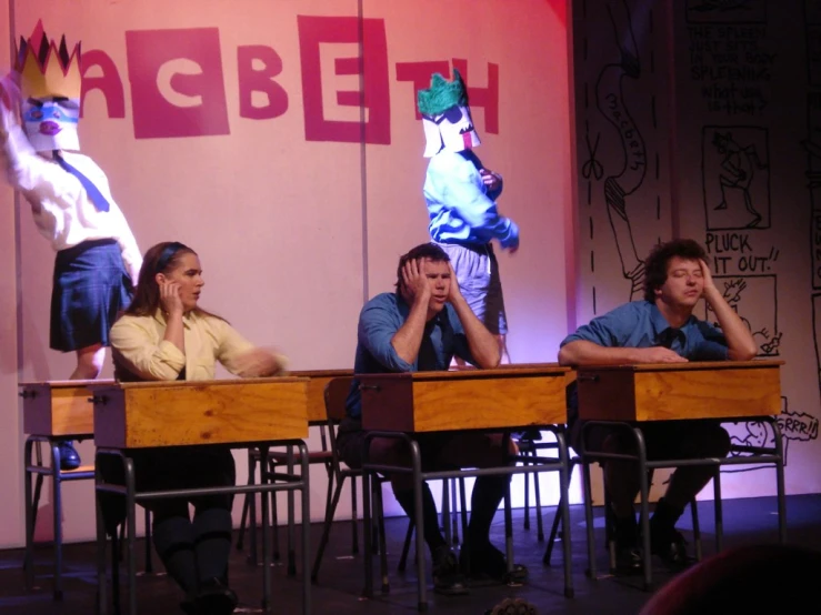 four people sit on a stage on wooden desks