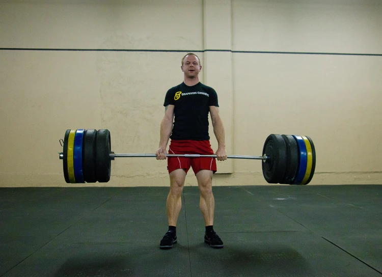 a man wearing red shorts, black shirt, and a black shirt, lifts a large metal barbell with two hands