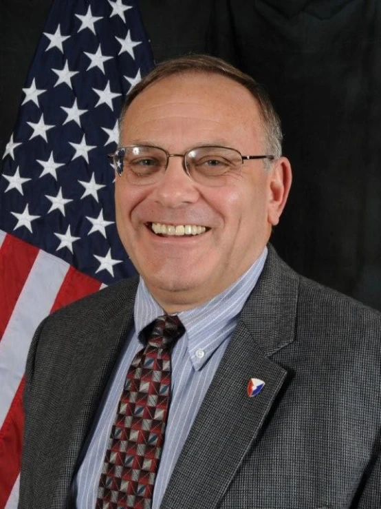 a man with glasses smiling in front of an american flag