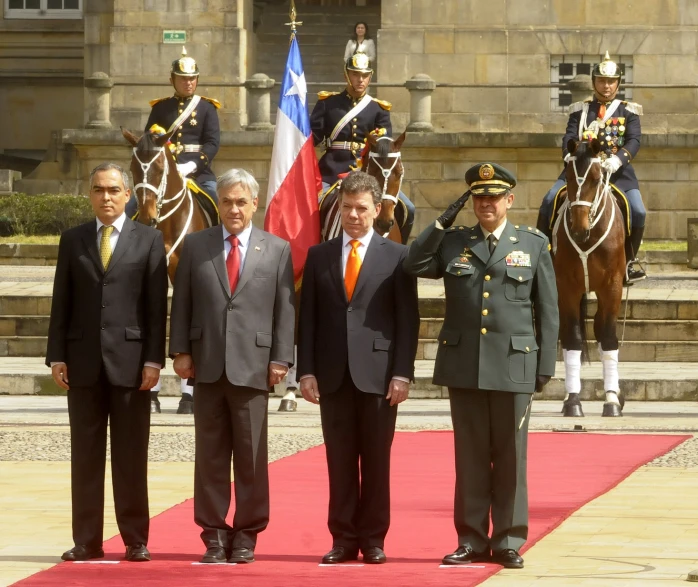 two men in suits standing by a flag, with military officers