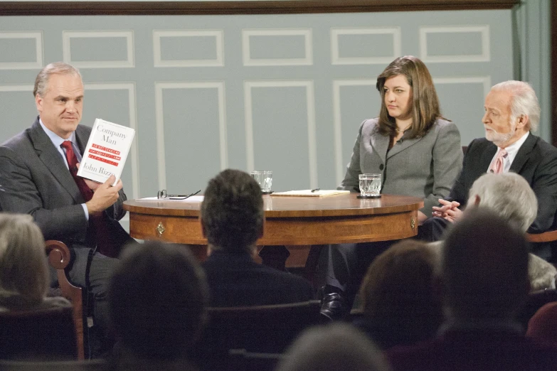 people sitting at a table with books while one man stands in front of the microphone