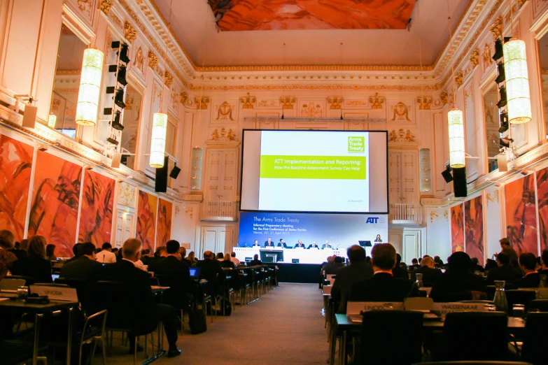 a conference hall filled with people in suits and sitting at desks, and projected screens displaying the audience