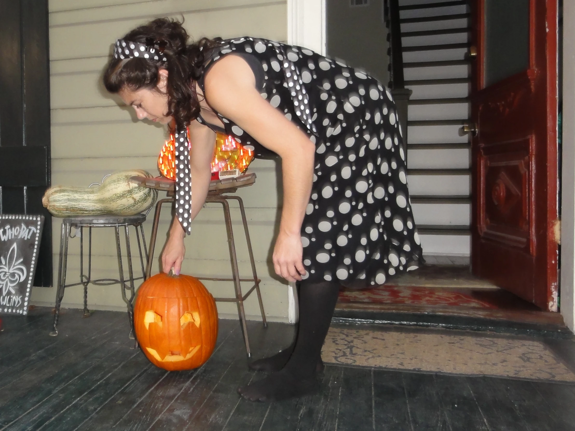 woman in black and white dress with black tights picking up pumpkin from porch