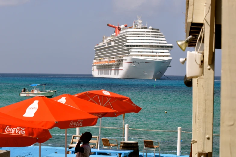 a cruise ship in the distance with an orange umbrella and chairs with red tables