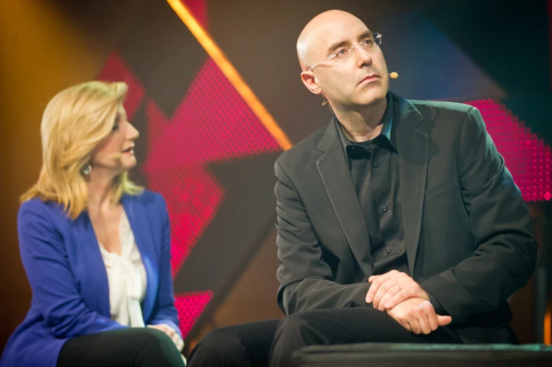 a man in a black jacket sitting on a chair next to a blonde woman